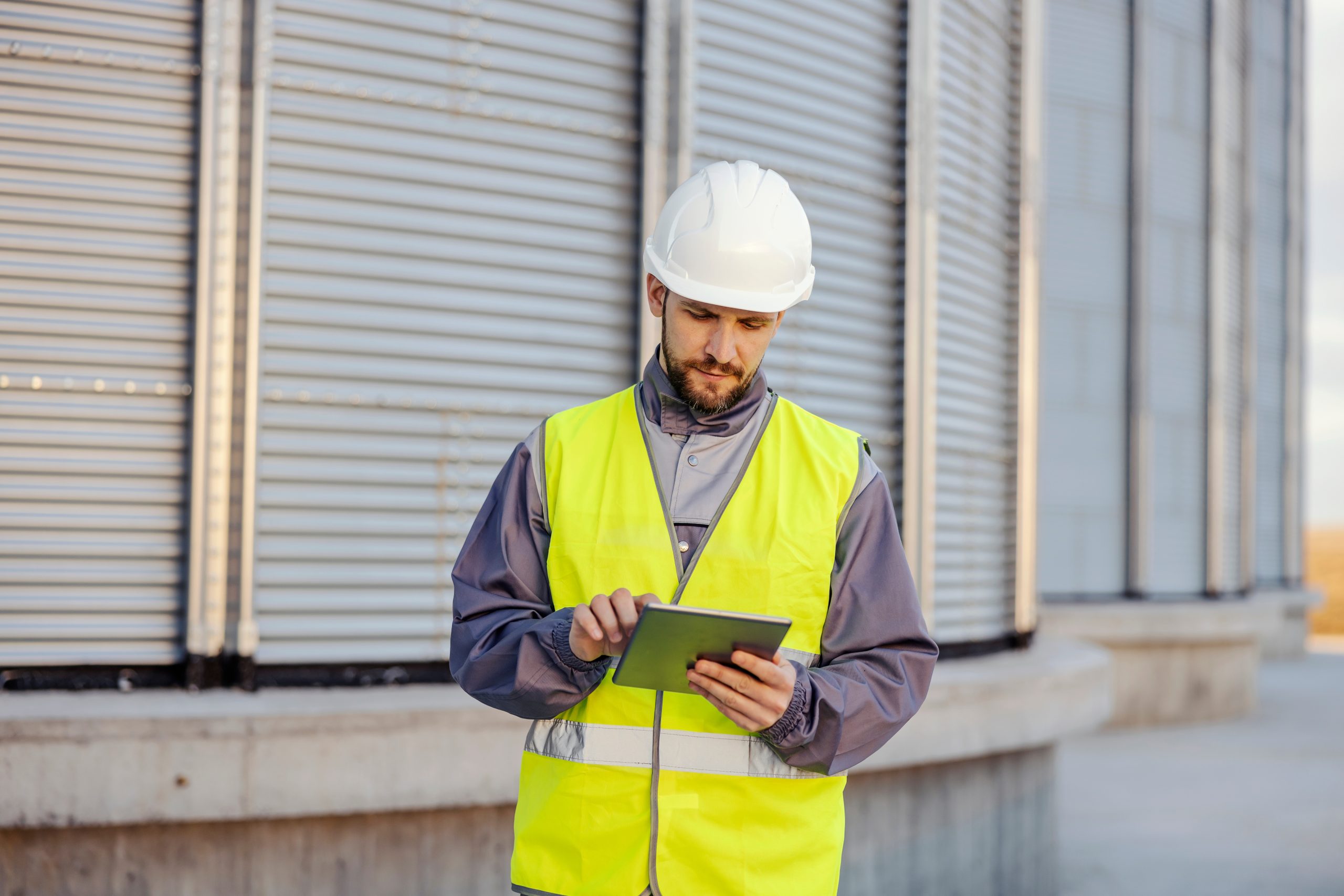 An industry worker is scrolling on tablet while standing next to a silos full of goods.